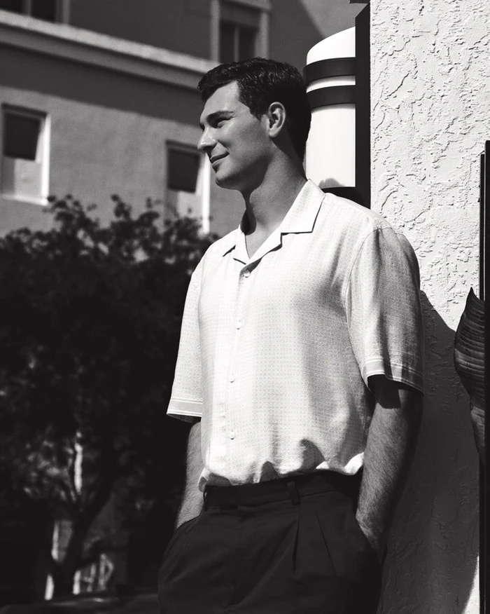 Black and white portrait of a man in a light short-sleeve collared shirt and dark pleated trousers standing outdoors.