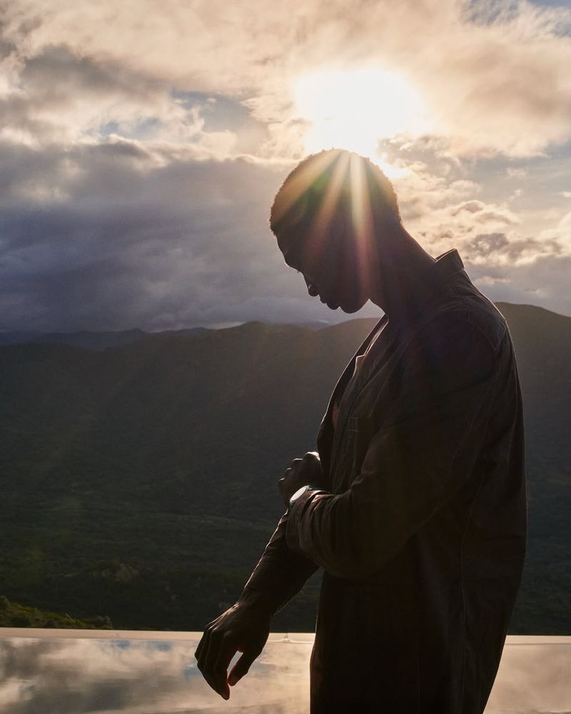 Silhouette of Black man rolling up sleeve, backlit by the sun, with a mountain landscape and cloudy sky in the background during the daytime.