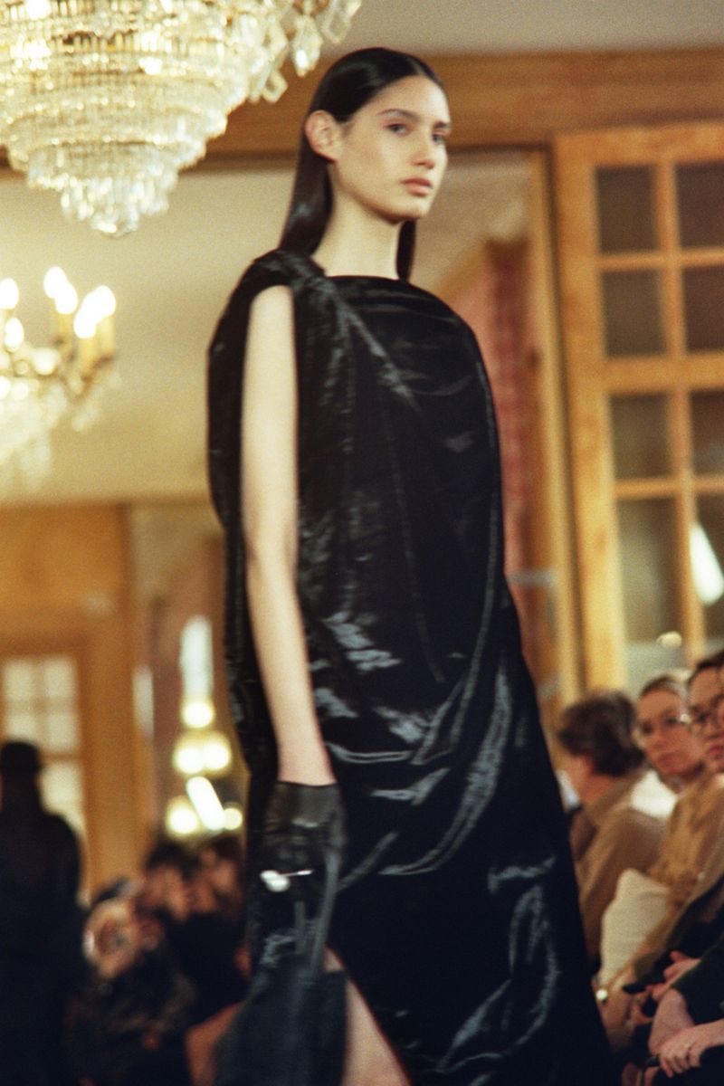 Runway fashion show with a model wearing a black dress and gloves, posing under a chandelier in an indoor event space with spectators.