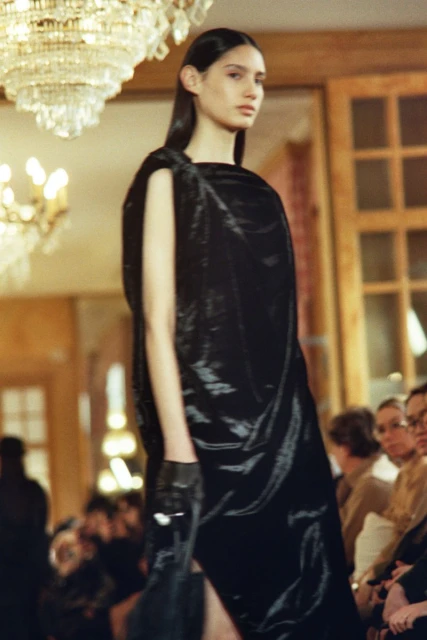 Runway fashion show with a model wearing a black dress and gloves, posing under a chandelier in an indoor event space with spectators.