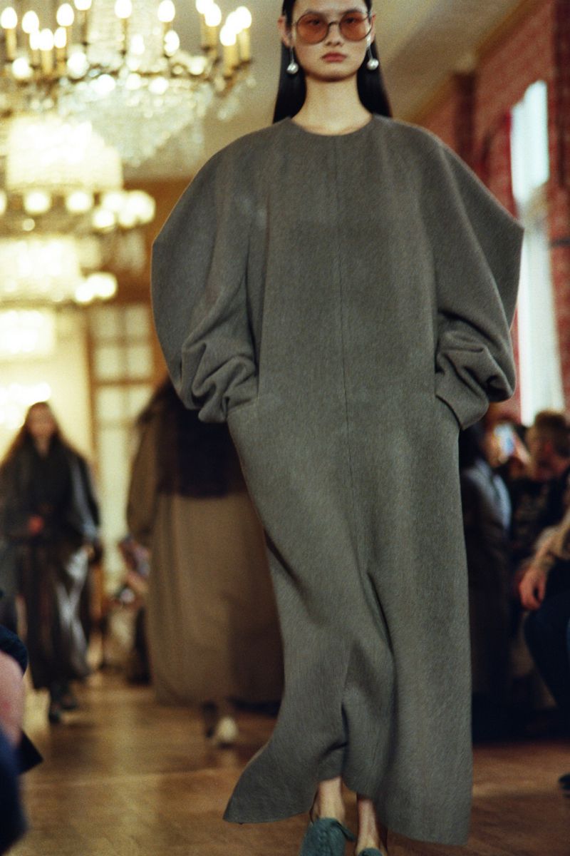 Runway shot of a model in a long gray coat with exaggerated sleeves, walking in an indoor space with warm lighting and an audience.