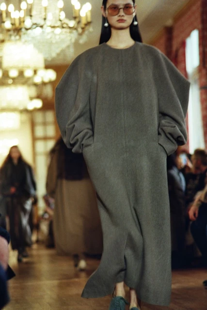 Runway shot of a model in a long gray coat with exaggerated sleeves, walking in an indoor space with warm lighting and an audience.