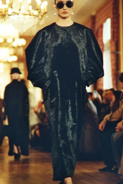 Runway model showcases a long, oversized black dress and large round sunglasses indoors, with audience members in the background.