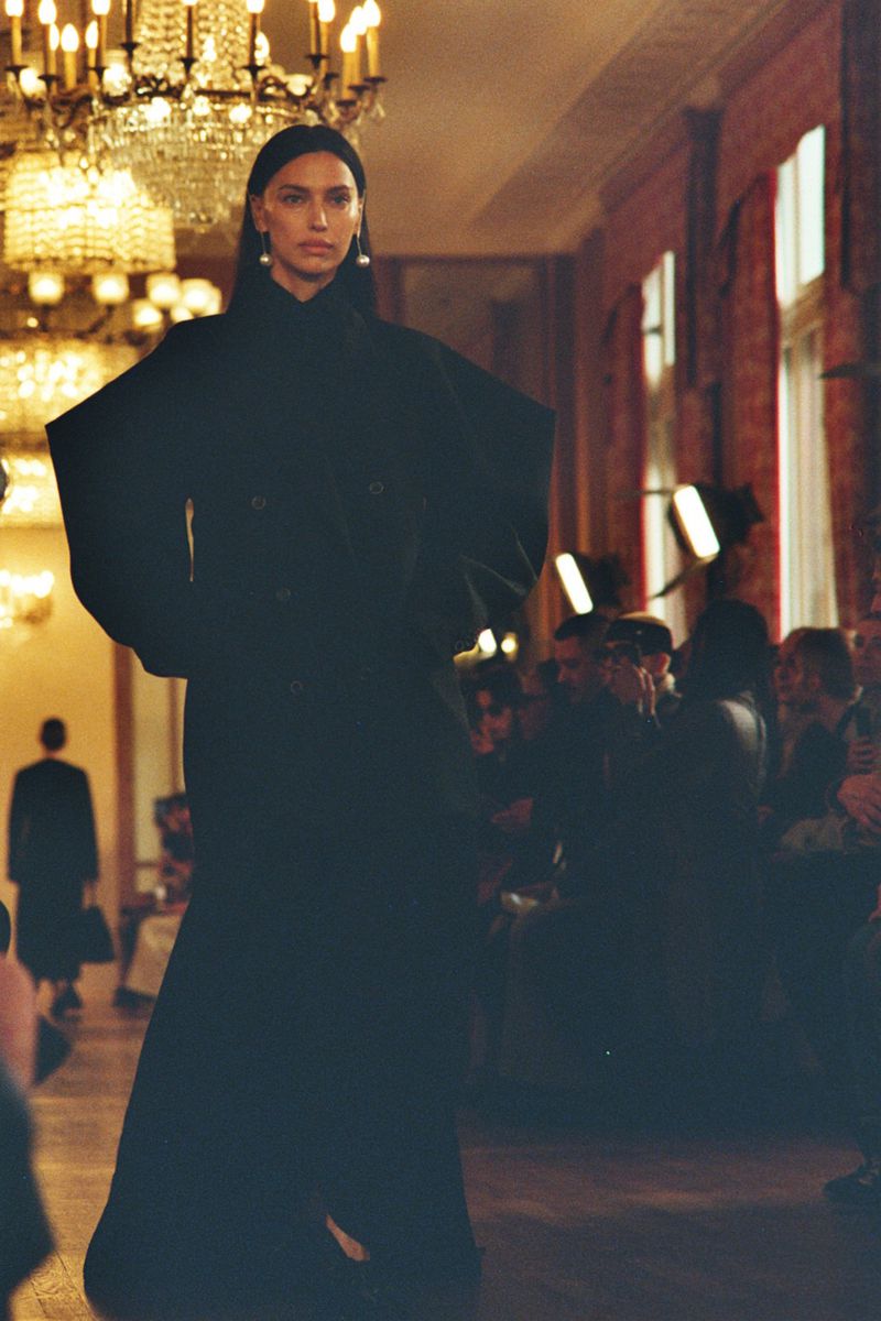 Runway show featuring a model in a black coat, chandelier, and crowd. The model has long dark hair and is wearing pearl earrings.