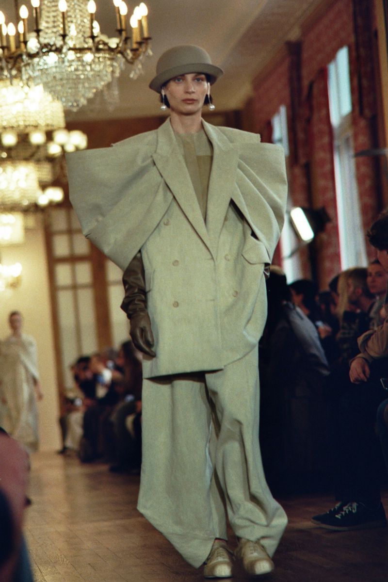 Fashion model on runway wearing an oversized light green pantsuit and a cloche hat at an indoor fashion show with an audience.