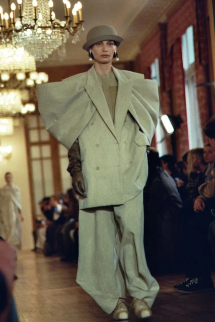 Fashion model on runway wearing an oversized light green pantsuit and a cloche hat at an indoor fashion show with an audience.