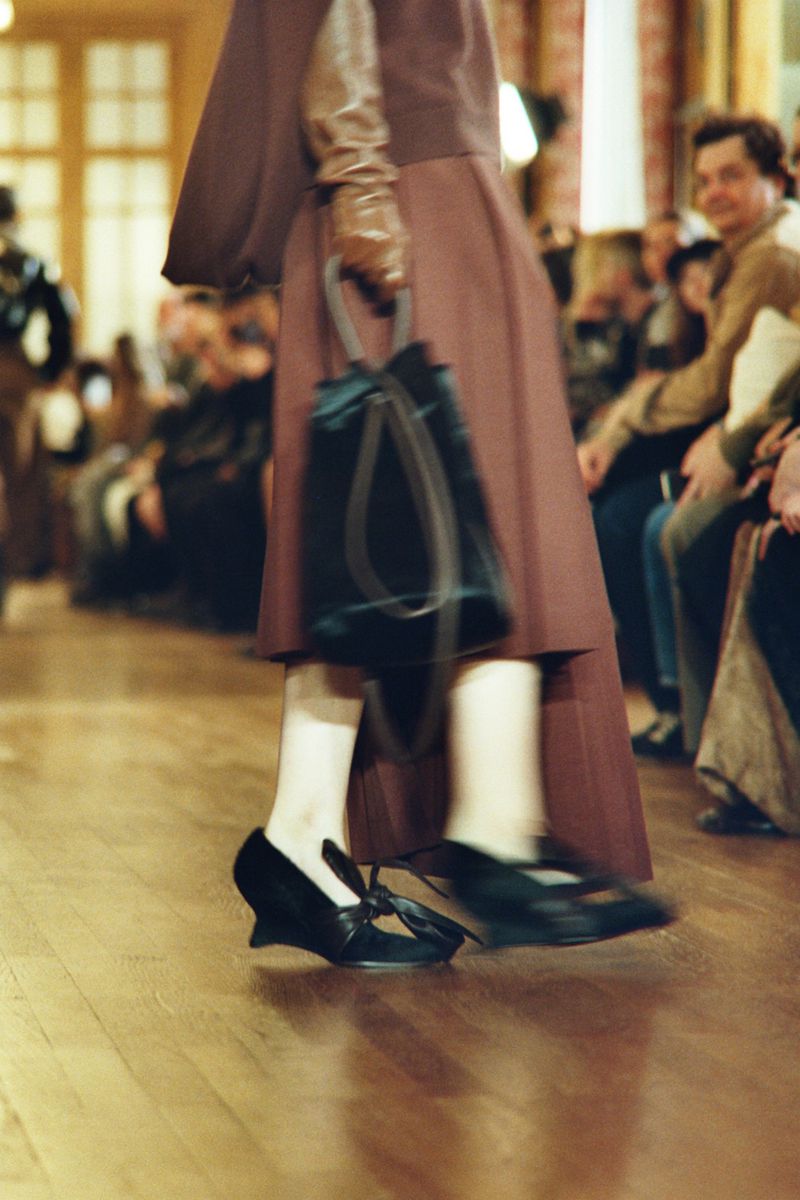 Runway fashion show: a model with blurred motion walking with a black bag, wearing a brown dress and black heels. Audience in the background.
