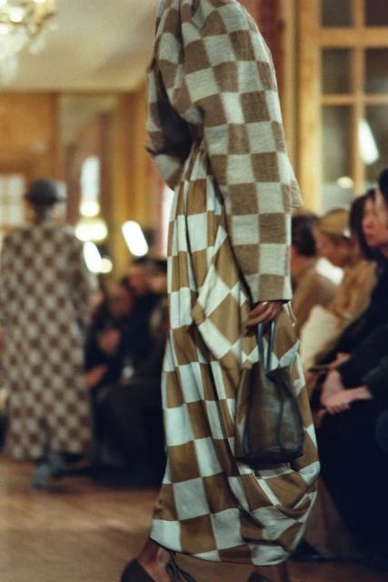 Runway fashion show featuring a model wearing a checkered coat and dress, holding a small purse, with audience members in the background.