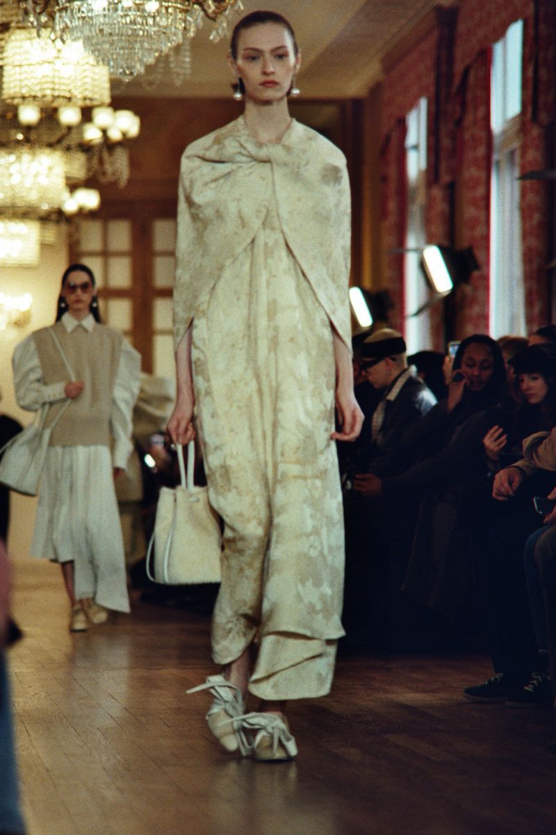 A model walks the runway in an ivory dress and cape with floral patterns, holding a white handbag during a fashion show indoors.