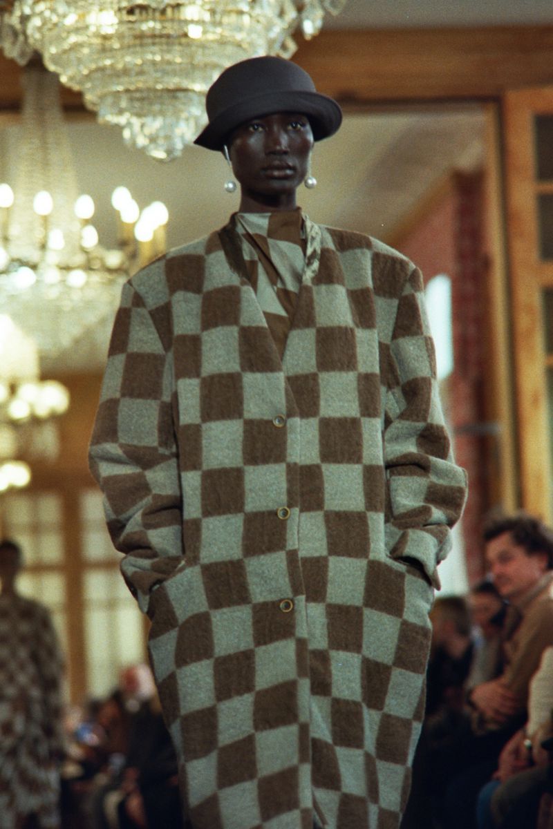 Runway fashion show featuring a Black female model in a checkered coat and bucket hat with a chandelier background and audience.