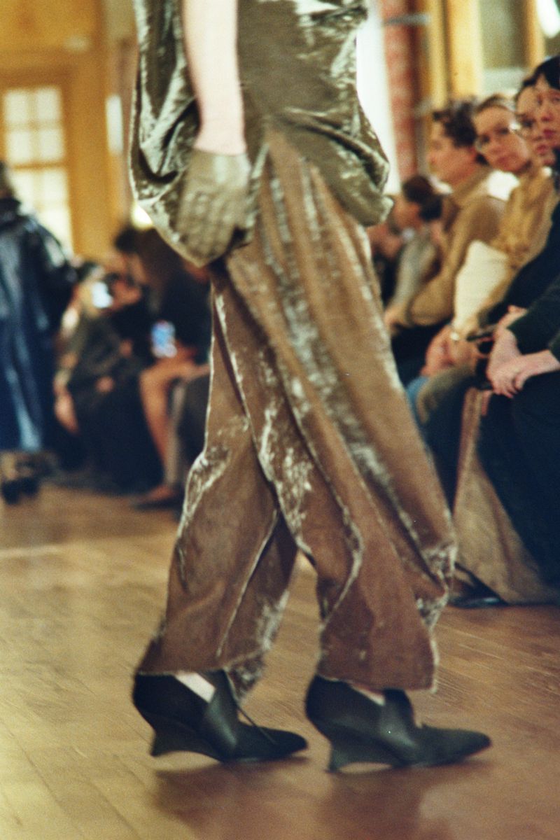 A fashion runway shot featuring a model walking, showcasing an olive green velvet dress and trousers with a crowd in the background.