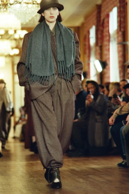 Runway model wearing brown suit and hat with gray scarf at indoor fashion show with audience in background, warm lighting and tone.