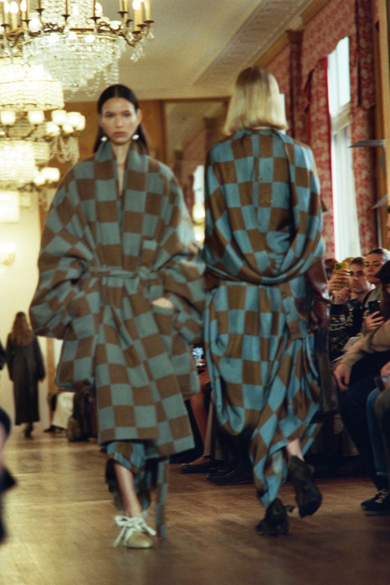 Two fashion models on a runway show off matching blue and brown checkered patterned outfits in an ornate interior with a crowd watching.