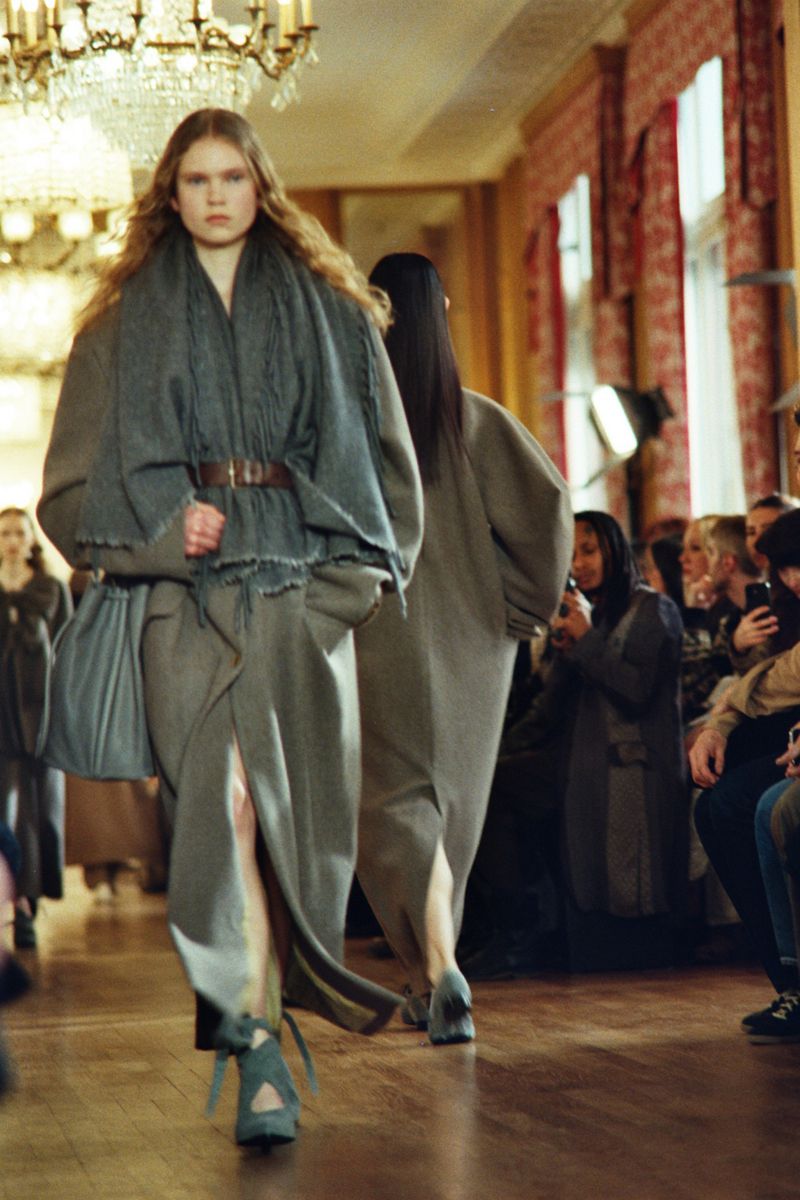 Runway fashion show featuring a woman in a grey draped outfit with belt and platform shoes, walking toward the camera in an indoor setting.