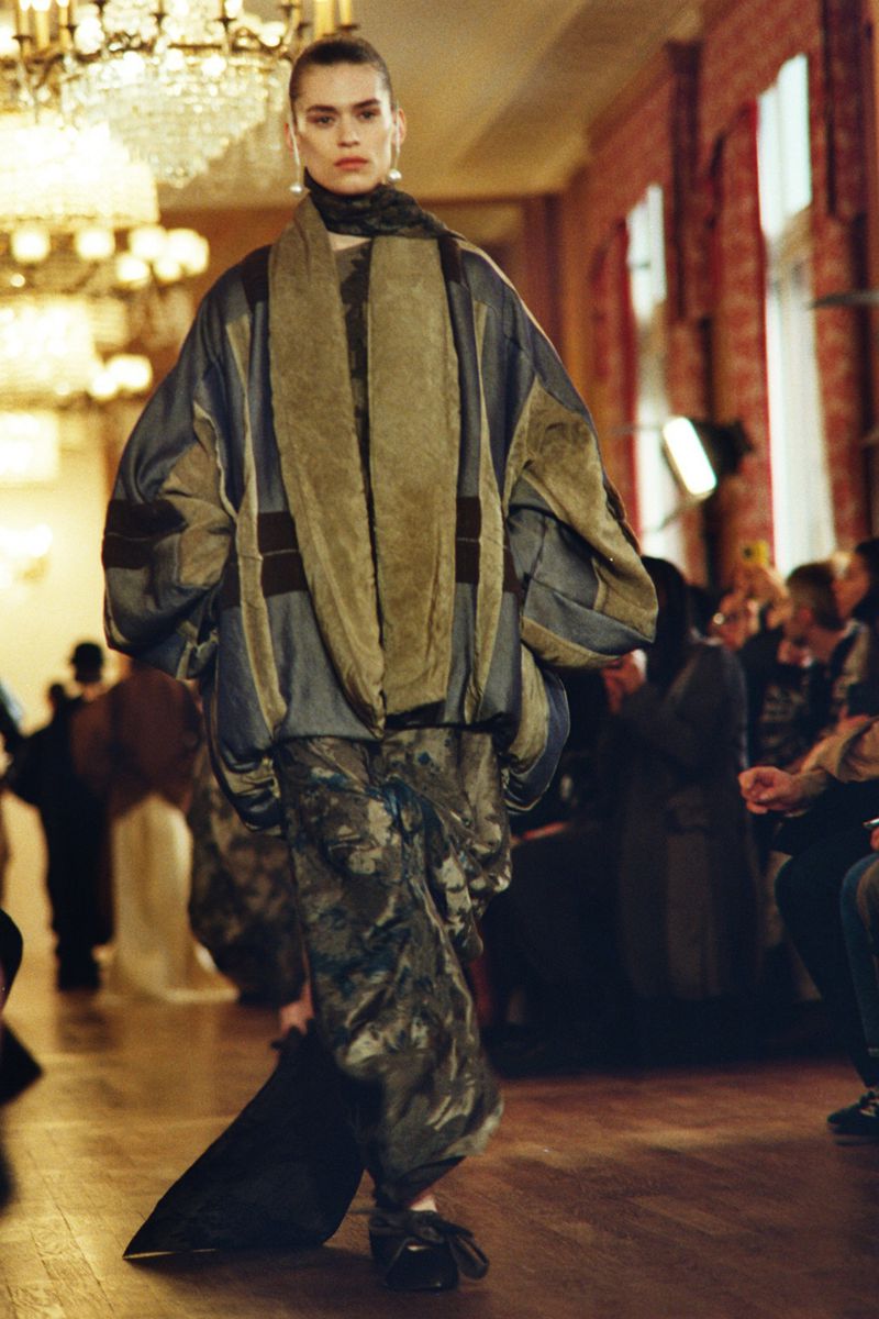 Runway fashion show featuring a model in an oversized patterned outfit, captured in a grand hall with audience members in the background.