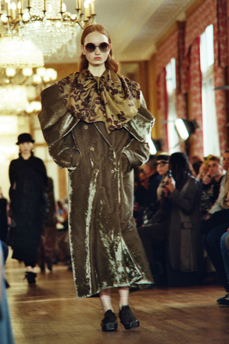 Runway model wearing a long olive-green coat with oversized shoulders and floral scarf, black shoes, and sunglasses, walking indoors.