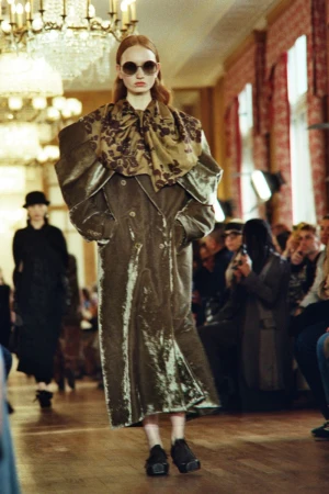 Runway model wearing a long olive-green coat with oversized shoulders and floral scarf, black shoes, and sunglasses, walking indoors.