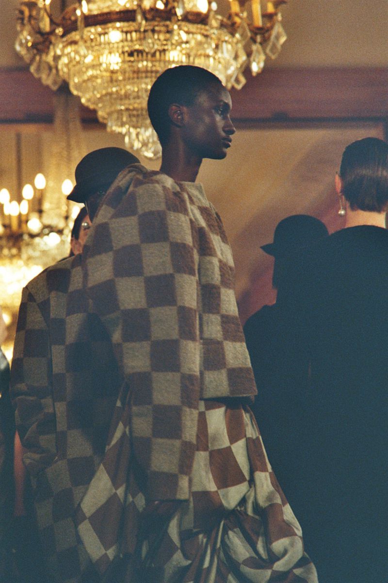 Runway fashion show featuring model in a checkered pattern dress with other attendees in the background and a chandelier overhead.