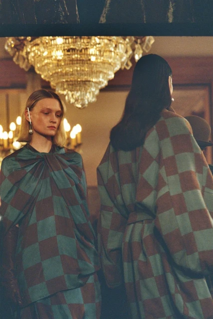 Two women in matching checkered dresses, one facing forward, the other with her back to the camera, in a room with a chandelier.