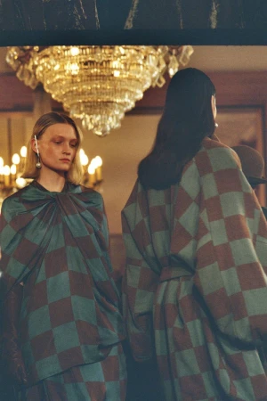 Two women in matching checkered dresses, one facing forward, the other with her back to the camera, in a room with a chandelier.