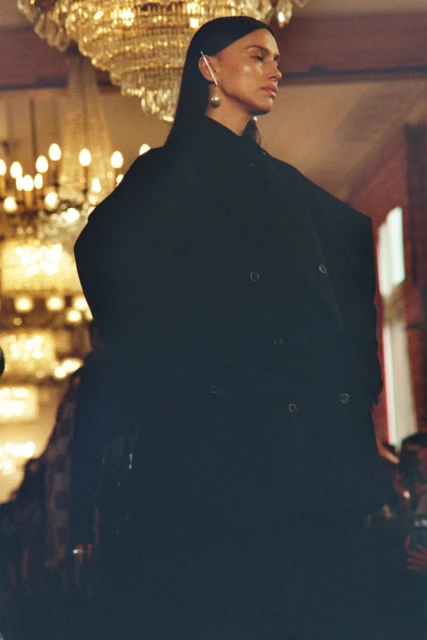 Woman in dark coat walks the runway at a fashion show with ornate chandelier lighting in the background and an audience watching.