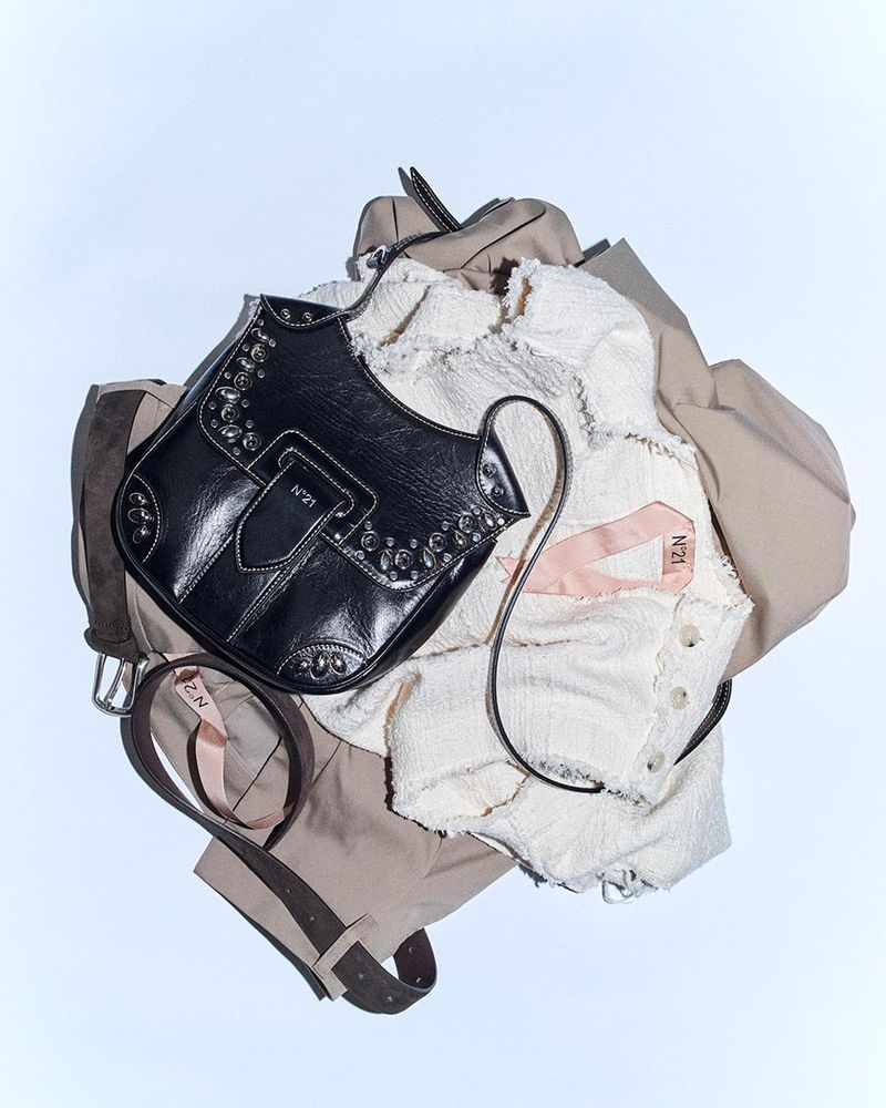 A black leather shoulder bag with metallic stud details sits atop a beige trench coat and white textured top on a white background.