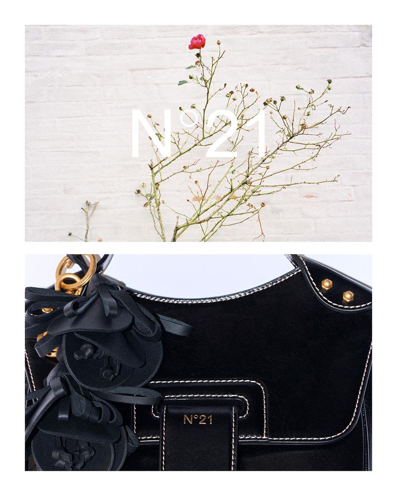 Studio shot of a black leather handbag with floral accents displayed against a textured white wall with a botanical accent and logo.