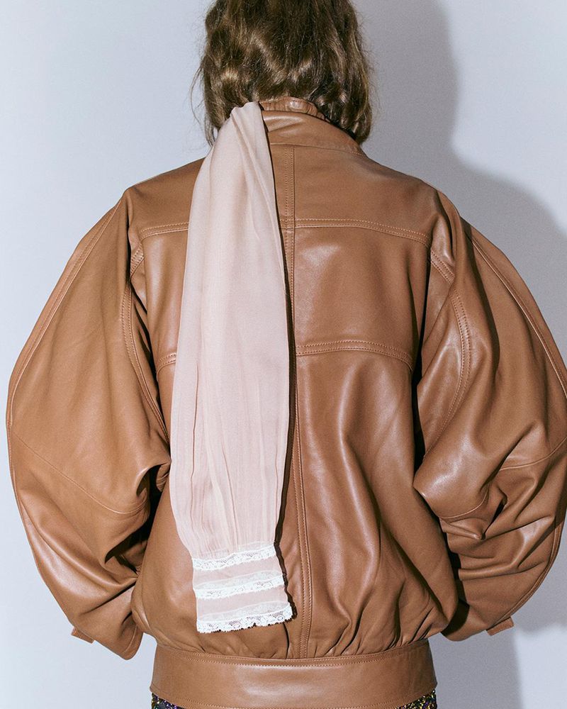 Back view of a woman wearing a brown leather jacket with oversized sleeves, paired with a peach top. Neutral indoor background.