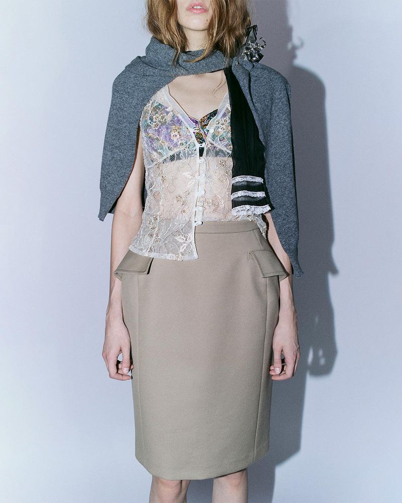 A woman poses in a beige pencil skirt, lace top, and gray cardigan, showcasing a fashion ensemble against a neutral background.