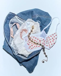Flat lay of blue denim, white ruffled underwear, red polka dot bralette, and a gold beaded necklace on a pale background.