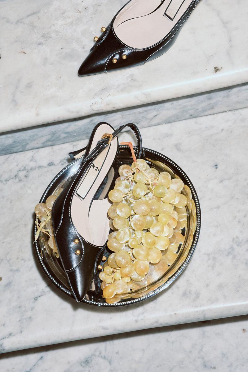 High-angle shot showcasing dark brown heels with gold accents on a silver tray filled with green grapes, set against a marble staircase backdrop.