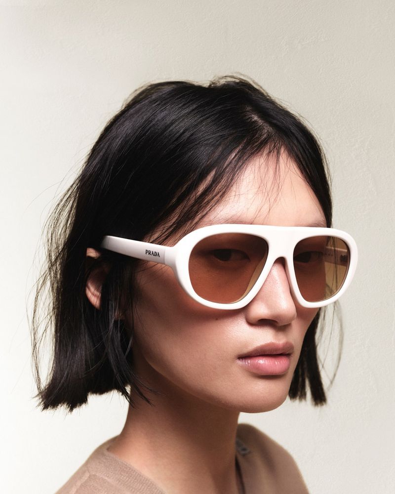 Close-up of a female model wearing white Prada aviator sunglasses with tan lenses against a neutral background in a studio shot.