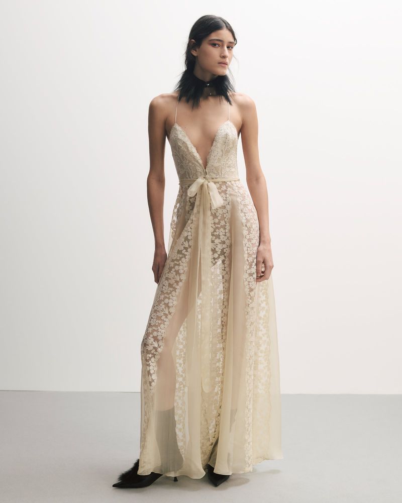 Full-length shot of a woman in a sheer, lacy gown with spaghetti straps and black heels, standing in front of a light gray background.