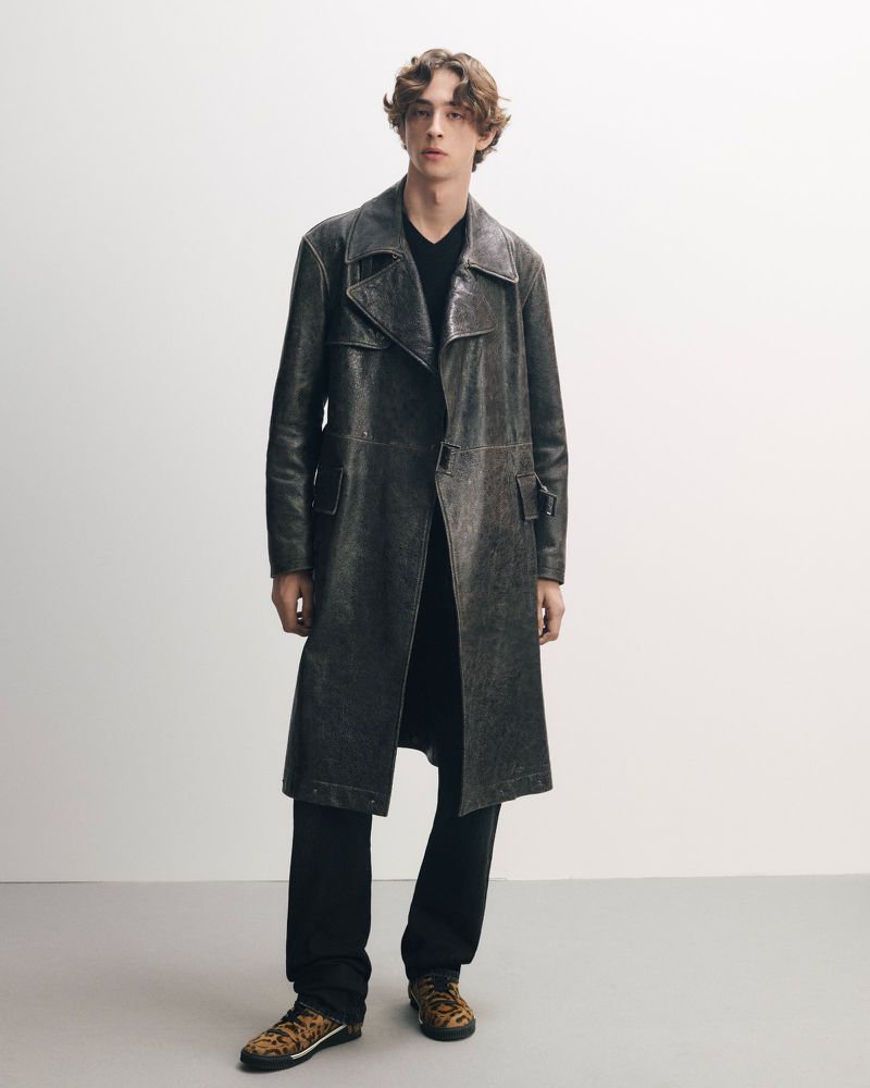 Full-length studio shot of a young adult male fashion model wearing an aged gray trench coat, black pants, and animal-print sneakers.