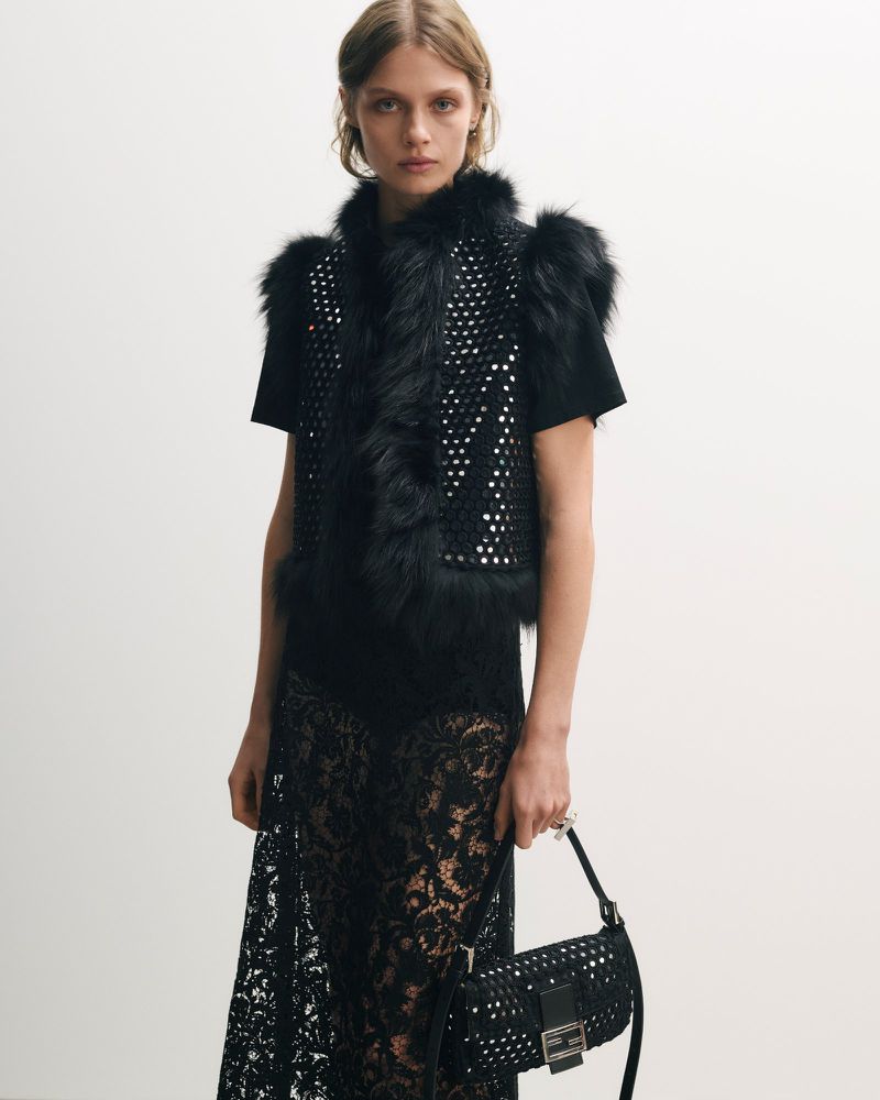Fashion shot of a model in a black fur vest and lace dress holding a handbag in a bright, neutral studio environment, looking at the camera.