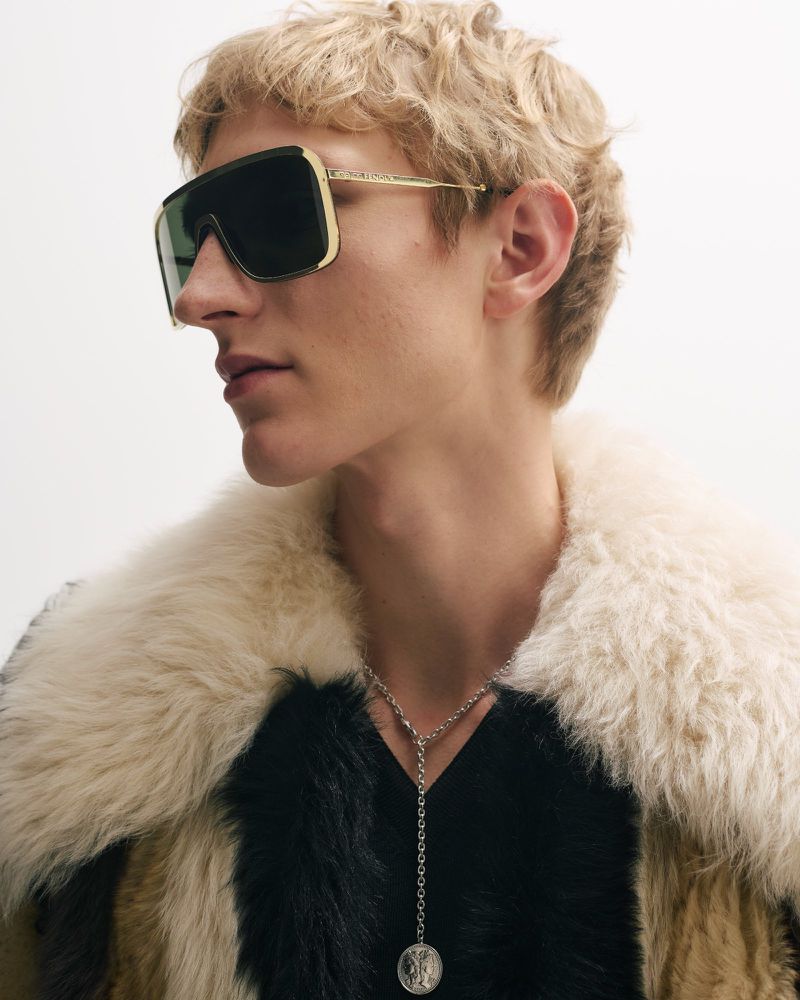 Adult male model posing in a studio wearing large sunglasses and a fur coat, showcasing fashion and style against a white backdrop.