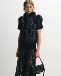 Model wearing a black studded fur-trimmed vest and sheer lace skirt while holding a black shoulder bag against a white background.