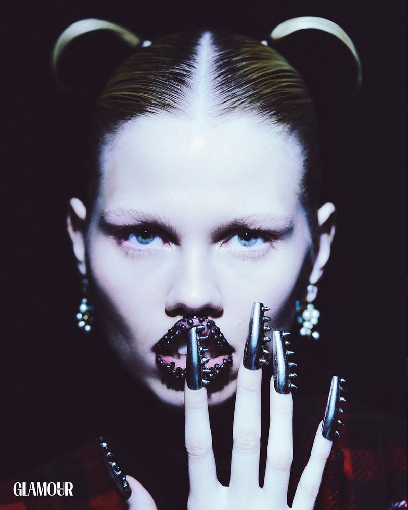 Close-up of a woman with unique metallic spike nail art and glossy lip jewelry against a dark backdrop for beauty or fashion.