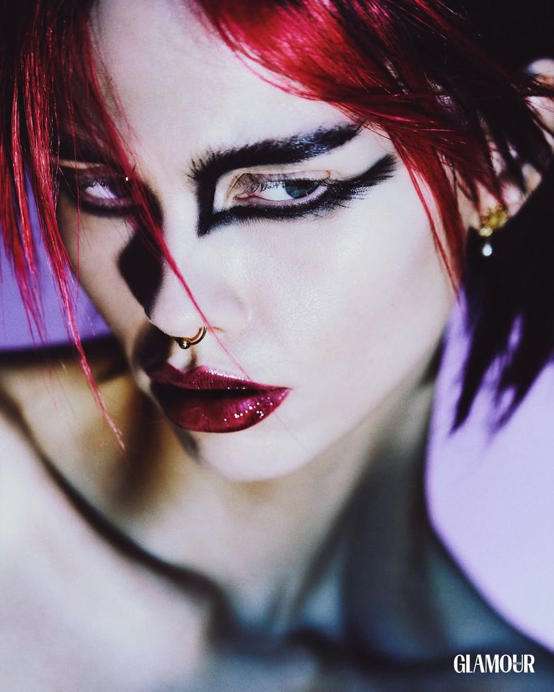 Close-up portrait of a fair-skinned woman with red hair, dramatic eyeliner, and glossy lips, posing against a blurred purple background.