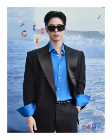 Man in a black suit and sunglasses posing against an abstract ocean-themed backdrop, fashionably dressed with a blue shirt.