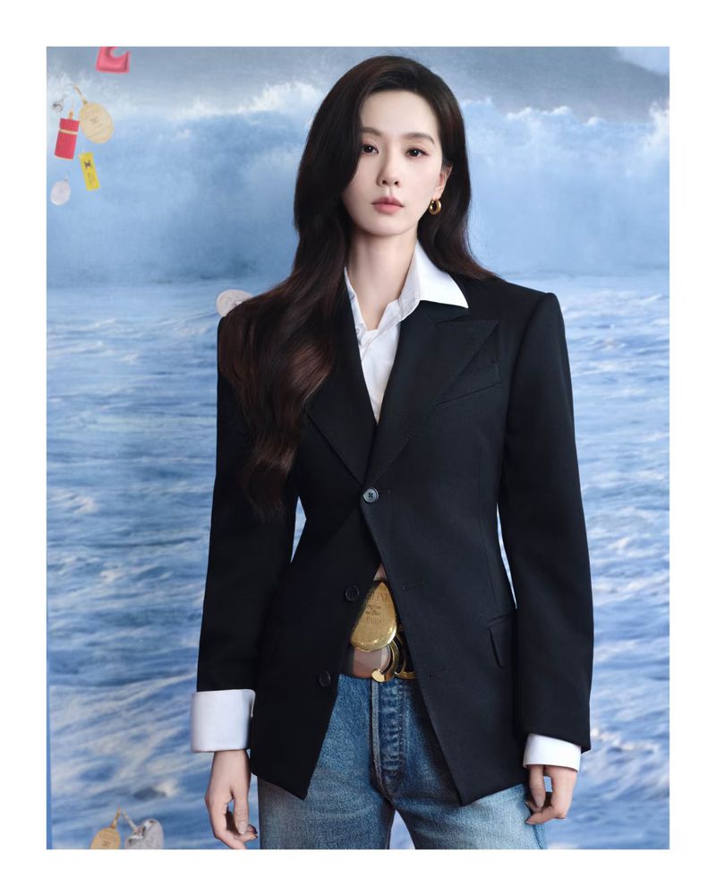 Portrait of an adult female with long, dark hair, wearing a dark blazer and jeans, set against a wavy ocean backdrop.