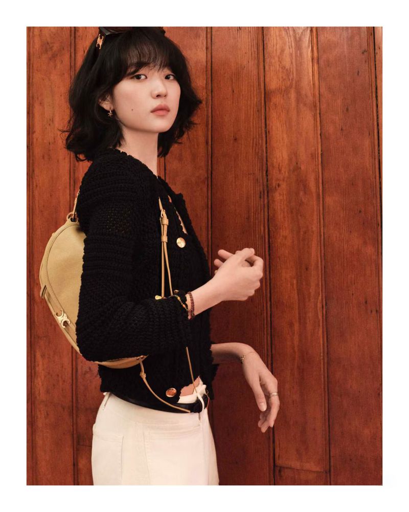 A young woman with short black hair poses indoors wearing a black jacket, beige purse, and white pants, near a wood paneled wall.