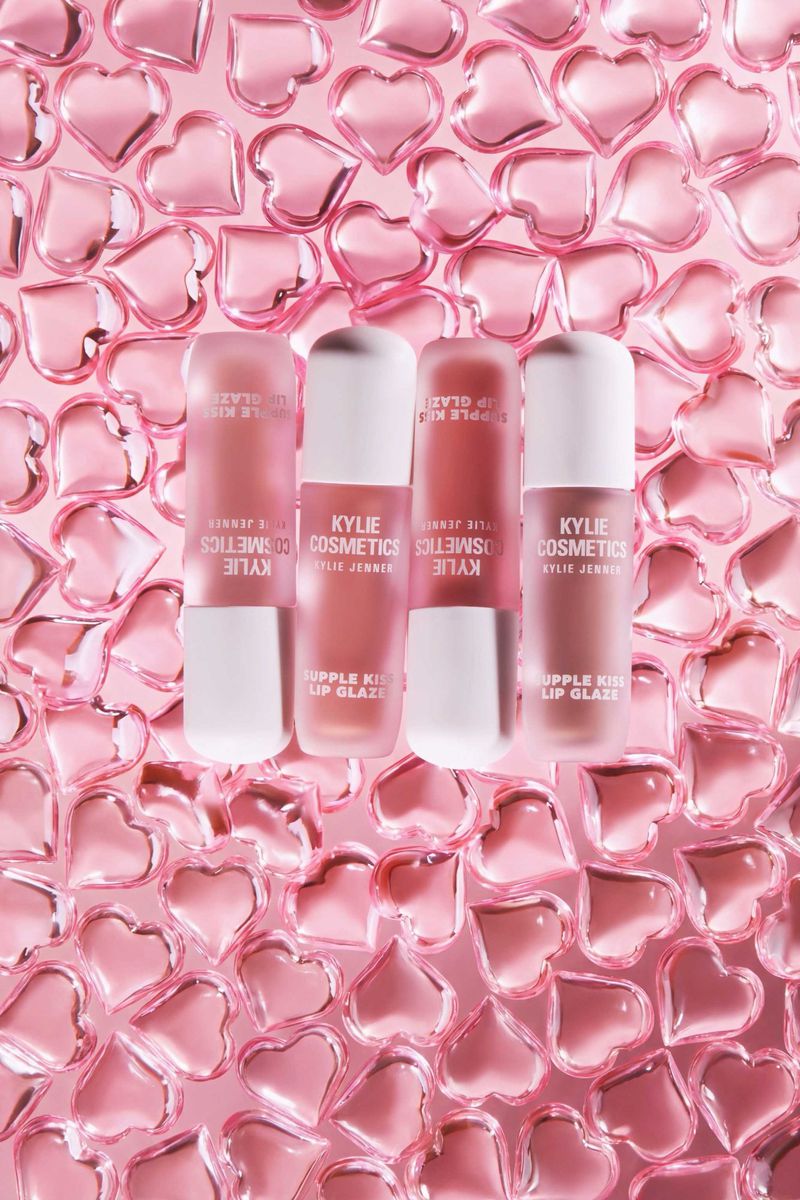 Kylie Cosmetics lip gloss products sit among many pink, translucent heart-shaped objects on a pink background in a studio shot.