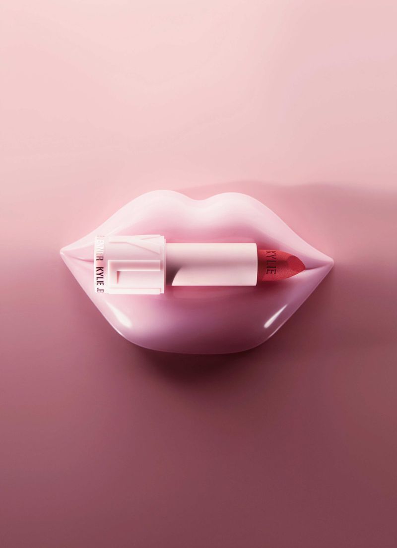 Close-up of lipstick tube held in glossy pink lips on pink background. The lipstick bullet is exposed, showcasing the vibrant red hue.