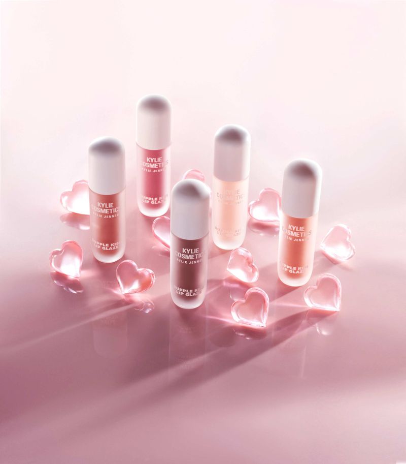 Five bottles of Kylie Cosmetics Simple Kind of Glaze lip gloss are arranged with transparent heart-shaped decorations on a pink surface.