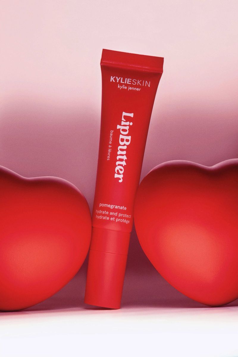 Kylie Skin Lip Butter tube in vibrant red, positioned between two red heart shapes against a soft gradient pink background. Beauty product.
