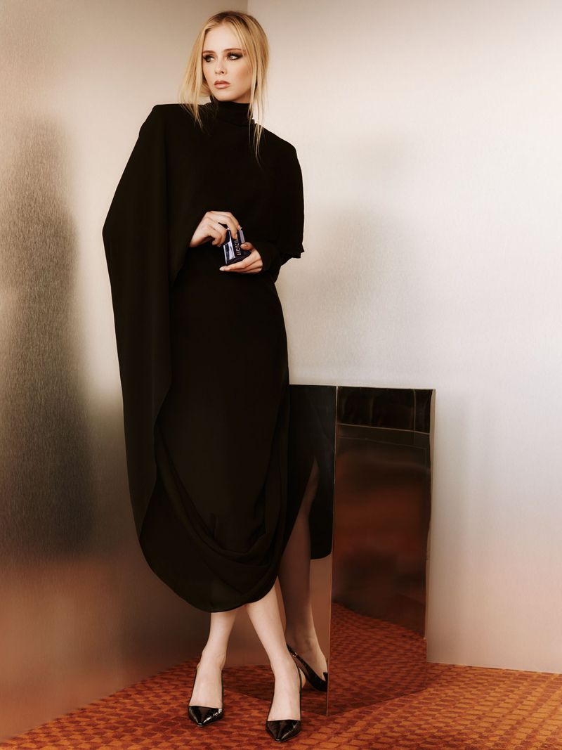 Elegant woman in black dress poses in studio with modern reflective surface, showcasing fashion and style in a sophisticated setting.