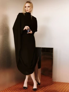 Blonde woman in a draped black high-neck gown holding a perfume bottle beside a mirrored plinth on patterned carpet.