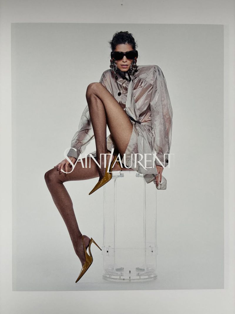 Fashion ad featuring a woman in sunglasses and hosiery, seated on a stool. She wears a jacket, and pointed-toe high-heel pumps.