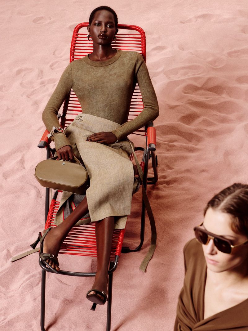 Dark-skinned woman in brown top and beige skirt sitting in a red chair on a sand dune, with another person visible in the foreground.